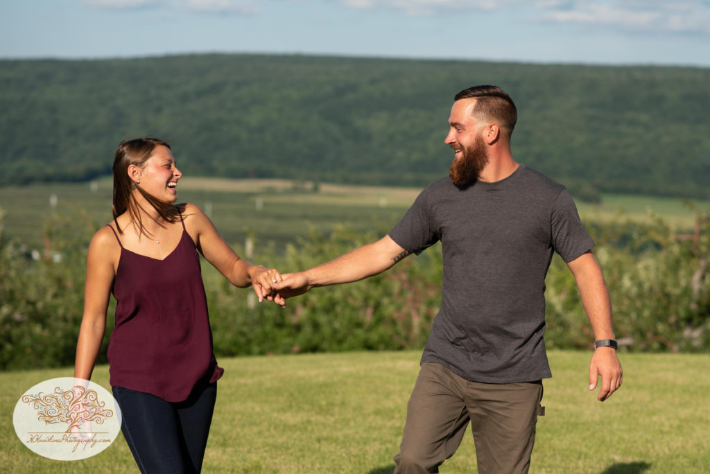 Engagement couple laughs together as they walk hand in hand surrounded by the hills of Lafayette NY and Route 20