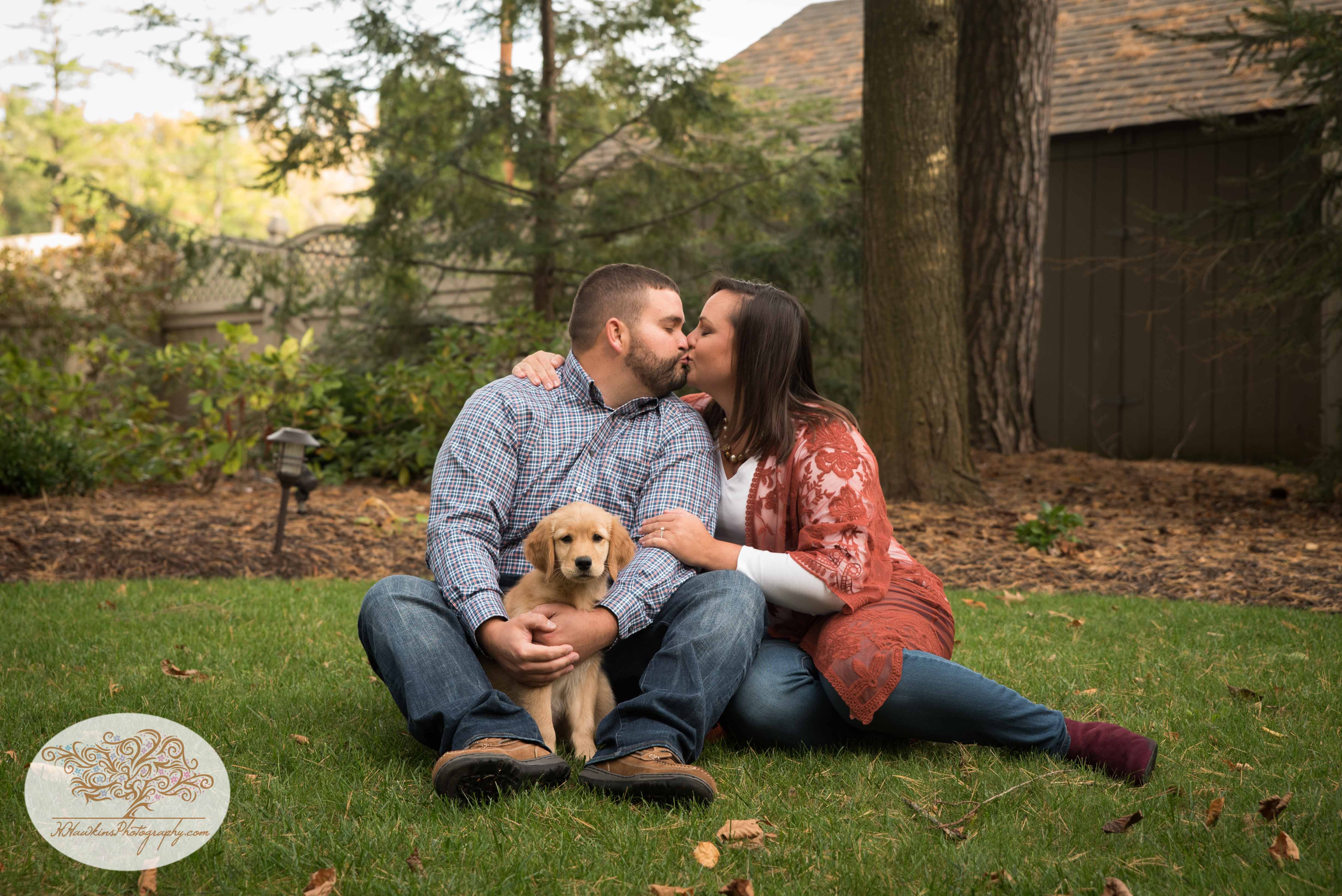 Groom kisses bride while golden retriever puppy sits in his lap during engagement photos