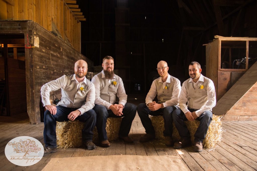 Groomsmen sit in upstate NY barn on hay bales for wedding portrait session