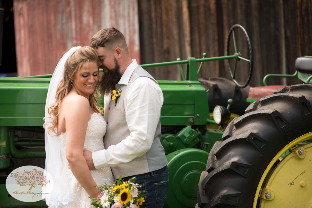 Bride smiles as groom nuzzles in front of John Deere tractor during Upstate NY barn wedding by Syracuse photographer