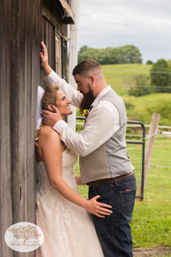 Bride leans against barn door as groom smiles at her during upstate ny farm wedding