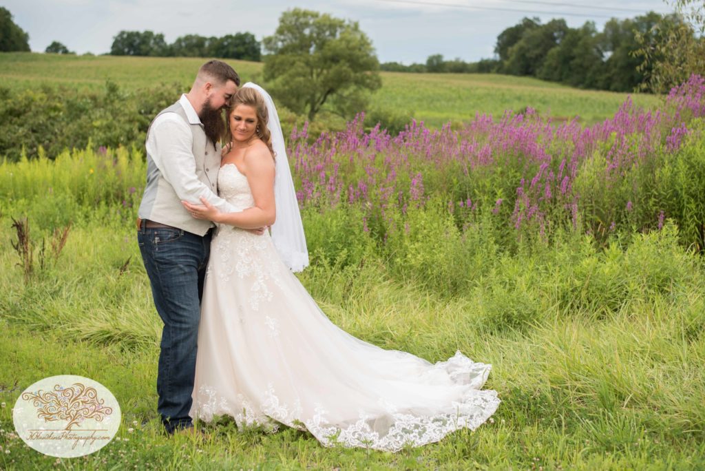 Bride and groom stand close together in farm field of purple wildflowers during Upstate NY barn wedding