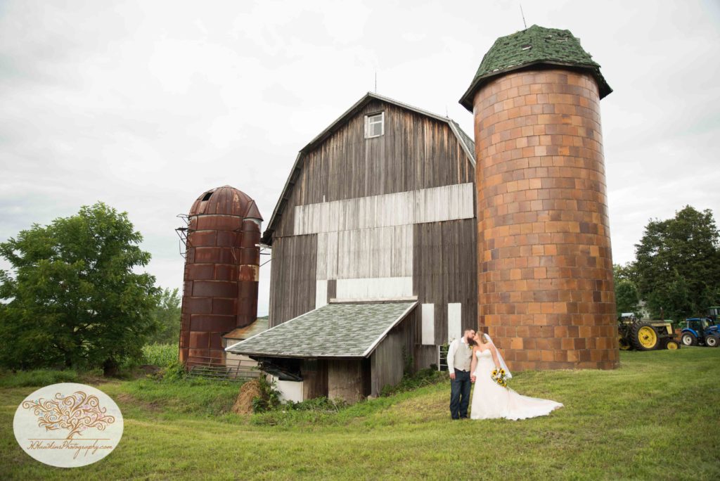 Bride and groom share a kiss in front of Upstate NY barn with two silos and tractors