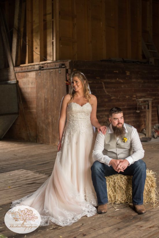 Bride stands next to groom sitting on hay bale in their barn in Upstate NY after wedding ceremony