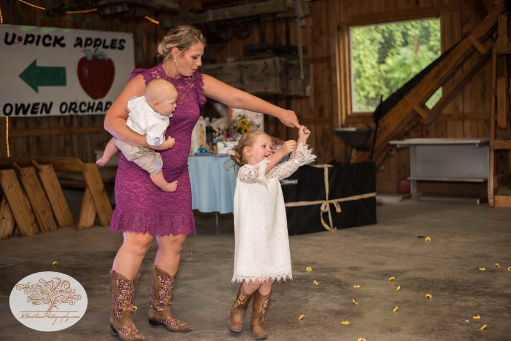 Mother spins flower girl while holding baby at Upstate NY barn wedding reception