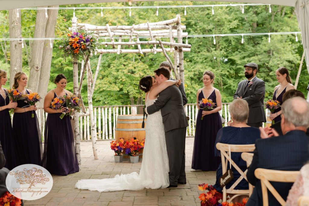 Bridal party looks on as bride and groom share a second kiss at the altar