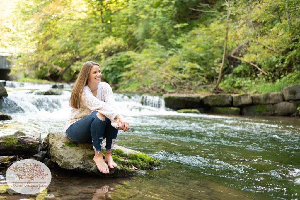 Senior girl sits on a rock on the side of a creek and smiles