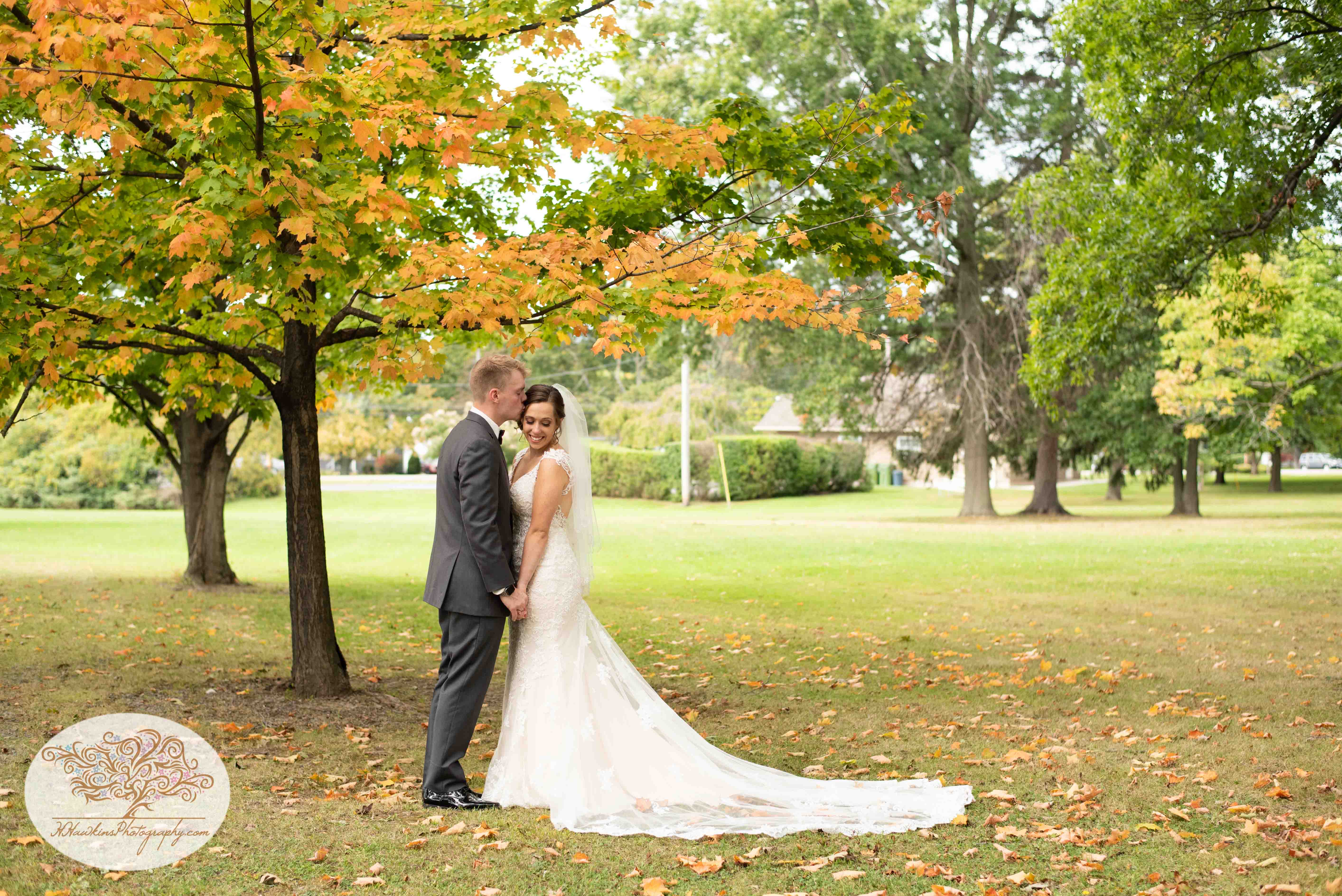 Groom kisses bride on the temple while she looks down standing in front of autumn tree