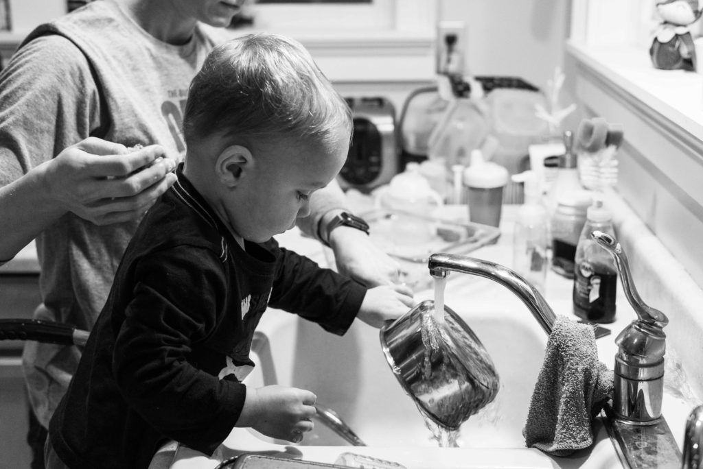 Little boy helps wash dishes and rinses a pan with water from faucet