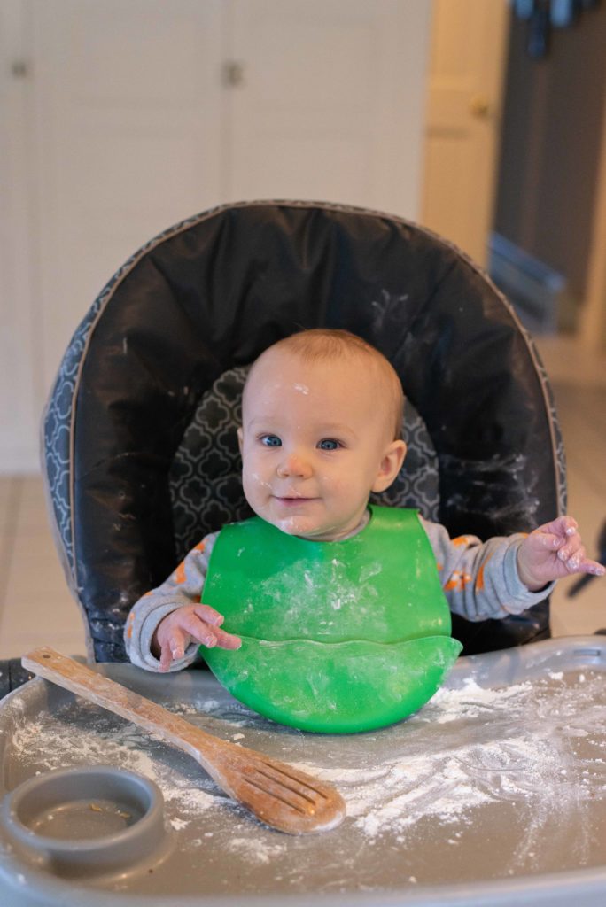 Baby sits in high chair with flour on his tray and wooden spoon at his hand