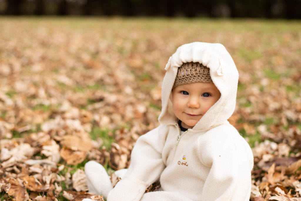 Baby boy in tan hat and white snowsuit sits on a lawn of fall leaves