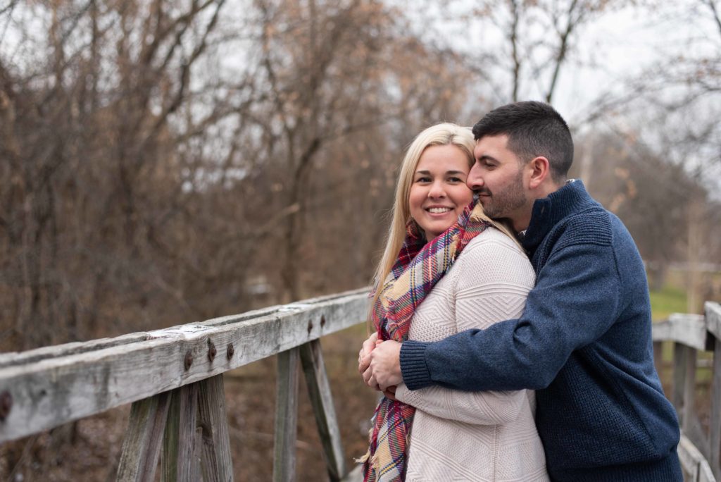 Groom wraps his arms around his bride and kisses her on the cheek as she smiles at the camera while standing on a wooden bridge of Camillus Erie Canal