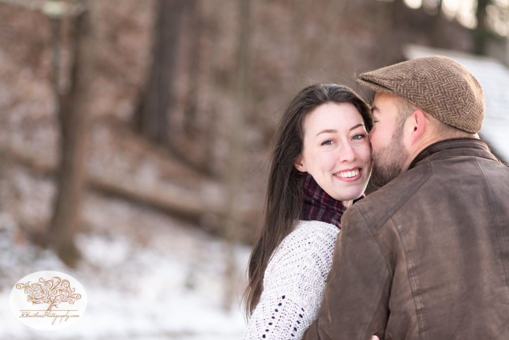 Groom kisses bride on the cheek as she looks over her shoulder during their snowy engagement pictures at Fillmore Glen