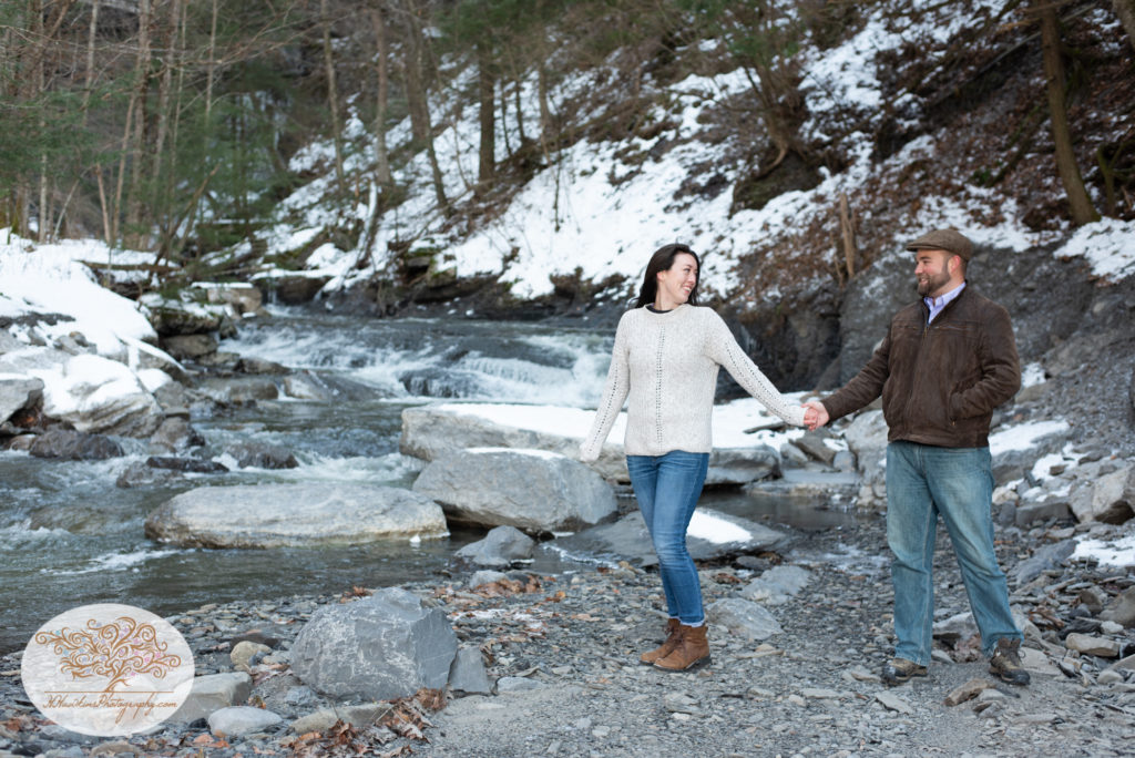 Guy and girl stand holding hands in front of stream at Fillmore Glen during their engagement session