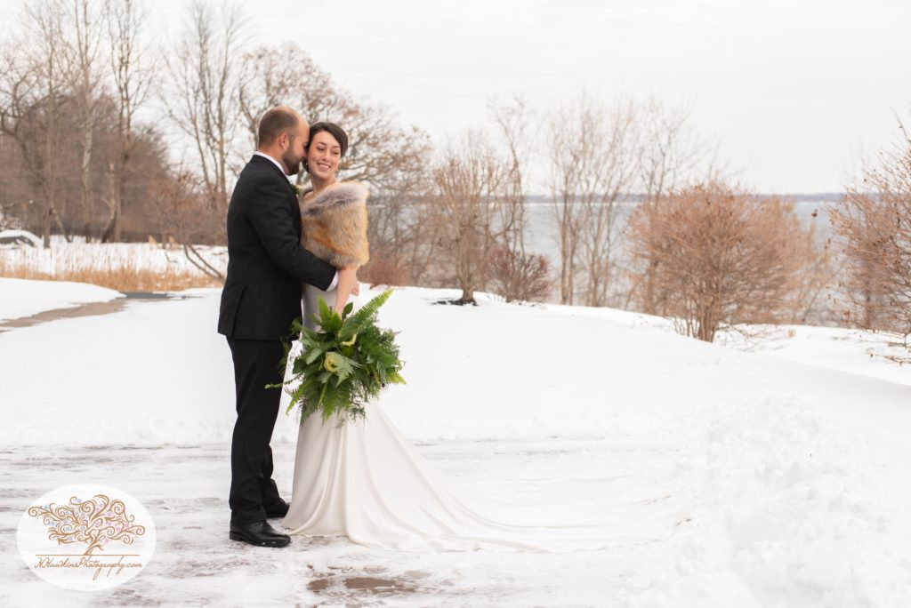 groom kisses bride on the cheek as she holds her green bouquet beside Seneca Lake