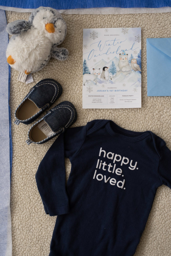 Penguin First Birthday Invitation spread with little boy shoes, onesie and stuffed penguin