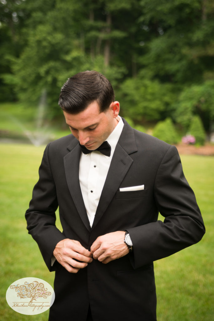 Groom buttons up his tux jacket outside in the yard before his wedding