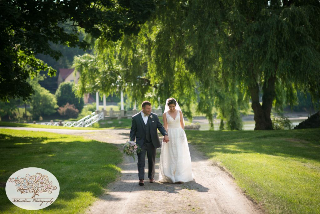 Bride and groom walk hand in hand on wedding day down a path at the park with weeping willow trees in the background