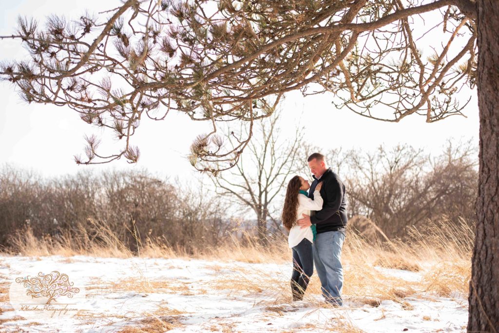 Engaged couple stand face to face framed by a pine tree in the snow with the sun behind them