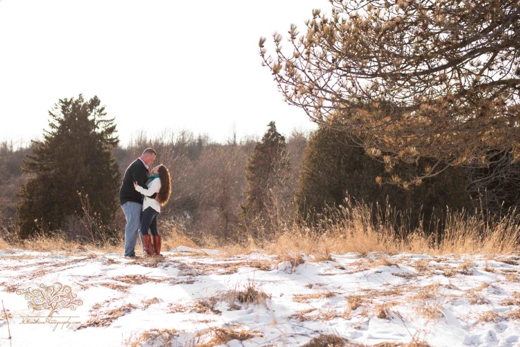 Syracuse wedding photographer captures engaged couple in a snowy sunny field with pine trees and tall grass