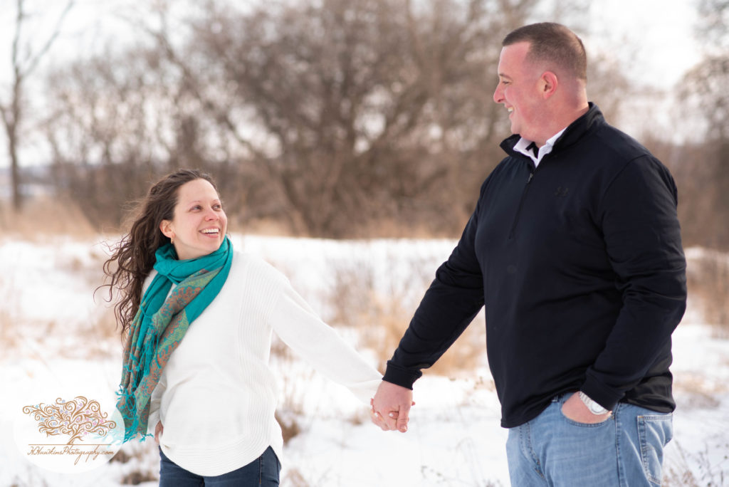 Bride to be looks at groom while holding his hand standing in a snowy field