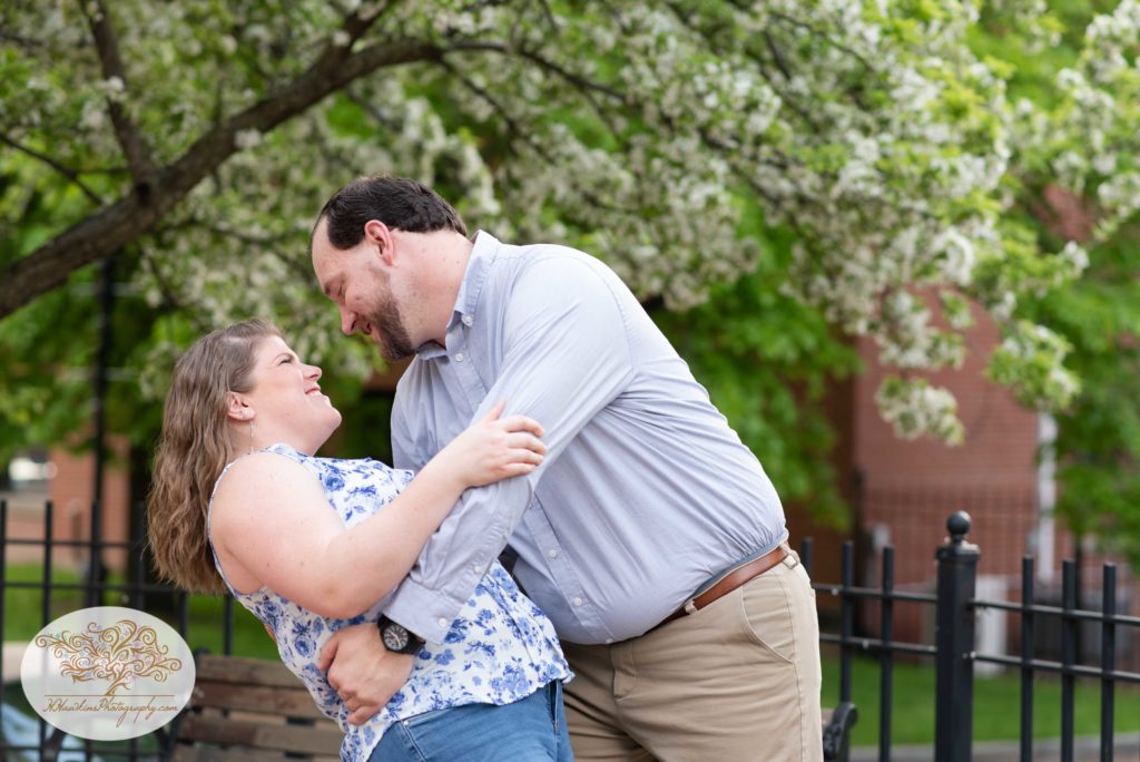 Groom dip kisses bride to be in front of a white blossoming spring tree at Syracuse's Franklin Square Park for their engagement pictures