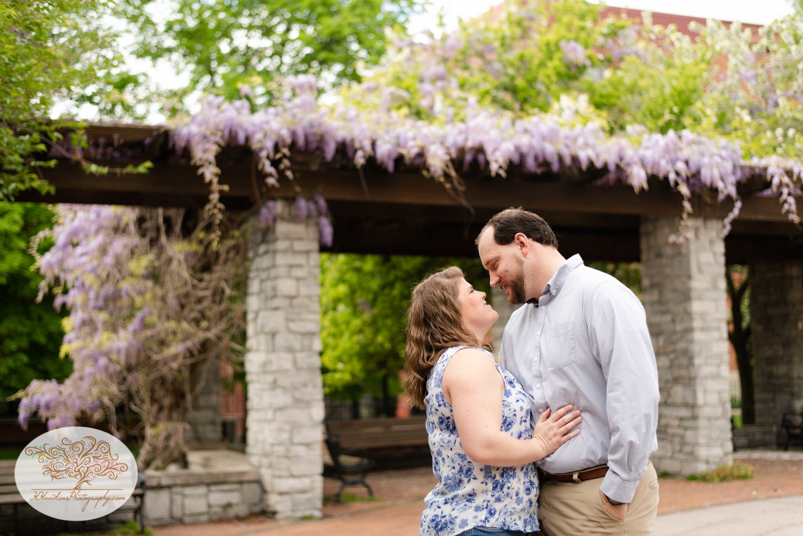Bride and groom to be stand in front of purple flowers and a pergola at Syracuse's Franklin Square Park