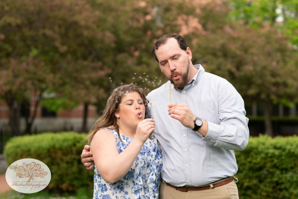 bride and groom to be blow dandelions gone to seed during their engagement picture session