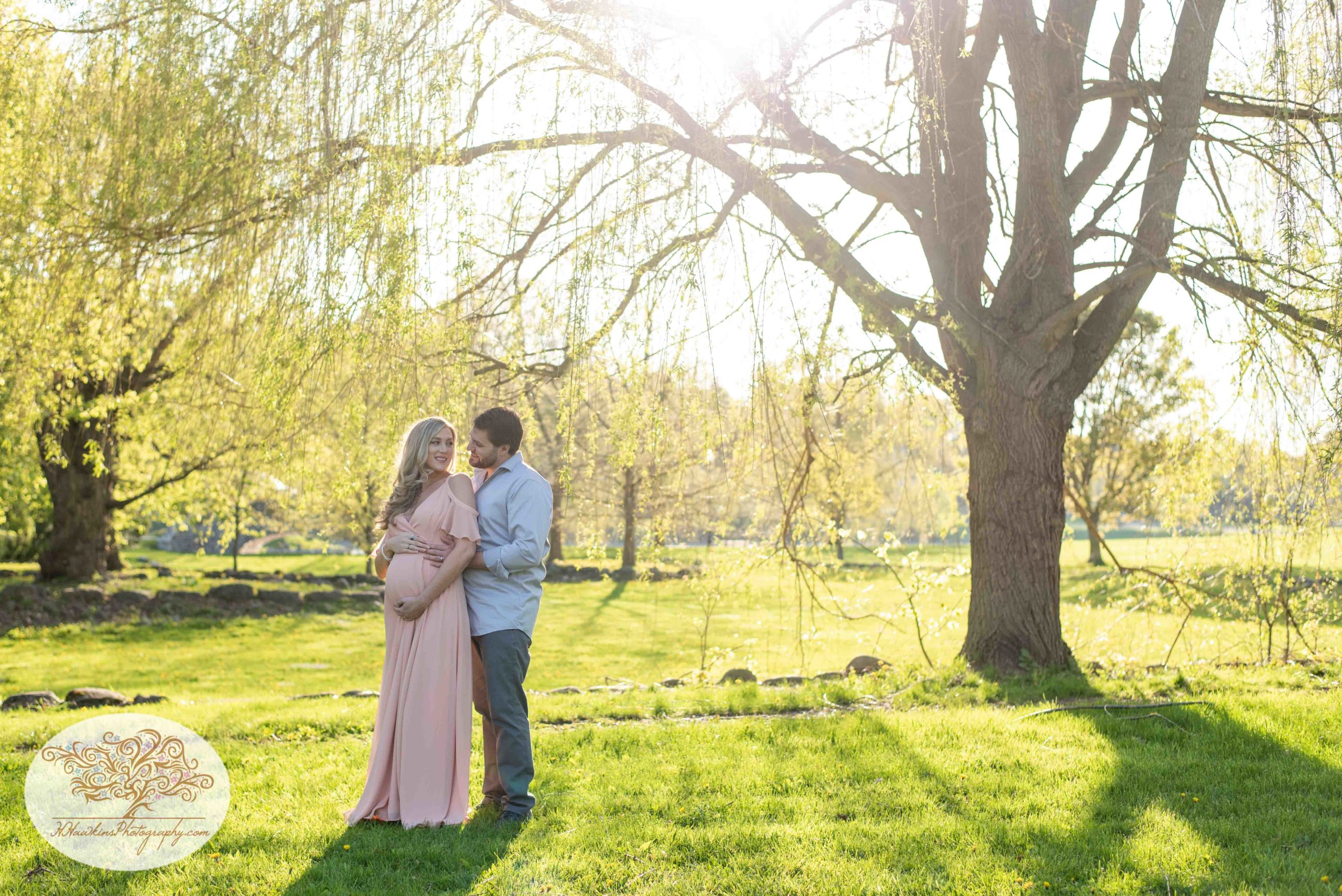 Expectant mom to be with her husband stands in front of weeping willow tree