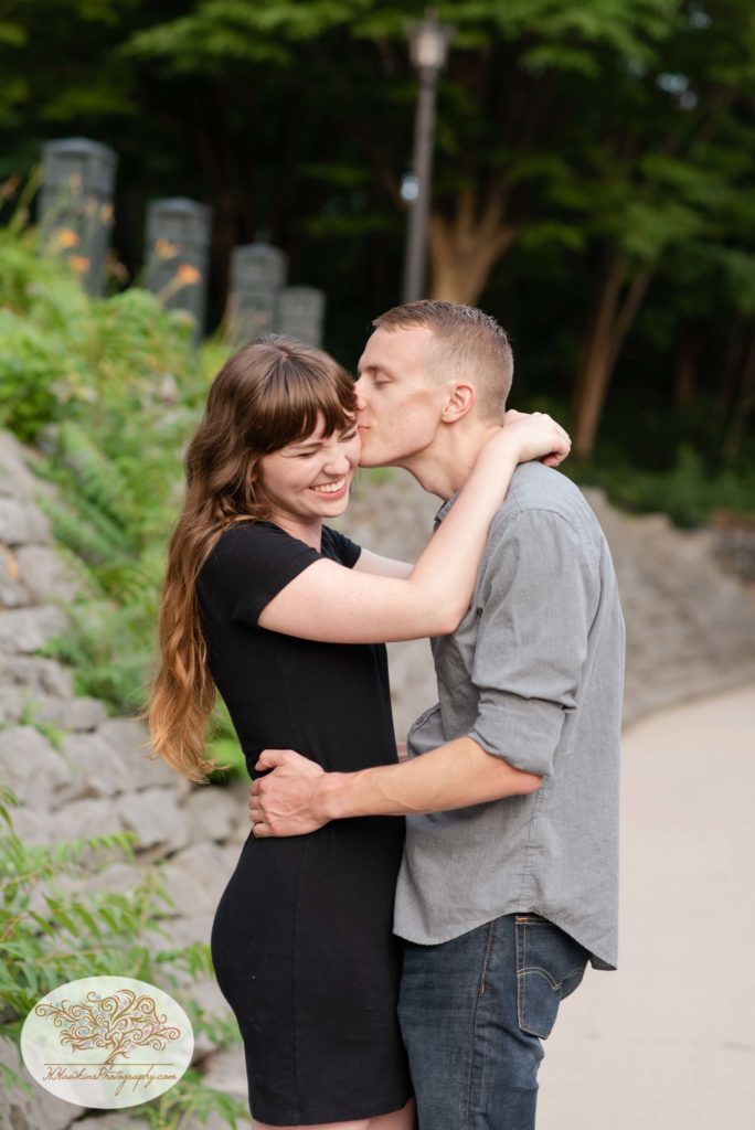 Groom kisses bride on cheek alongside a stone wall at Syracuse's Creekwalk