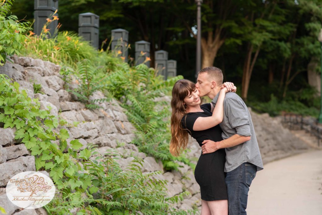 Groom kisses bride on the cheeck as they stand alongside a stone wall at Syracuse's Creekwalk