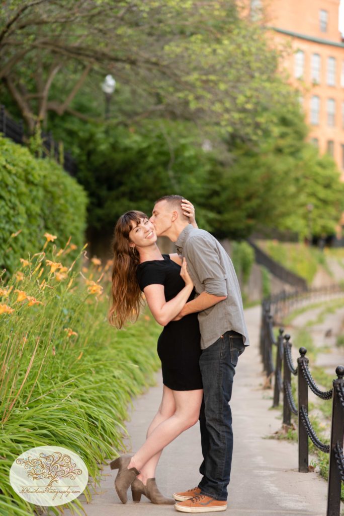 Bride to be puts her hand behind groom's head as he kisses her cheek alongside the stone wall on Syracuse's Creekwalk