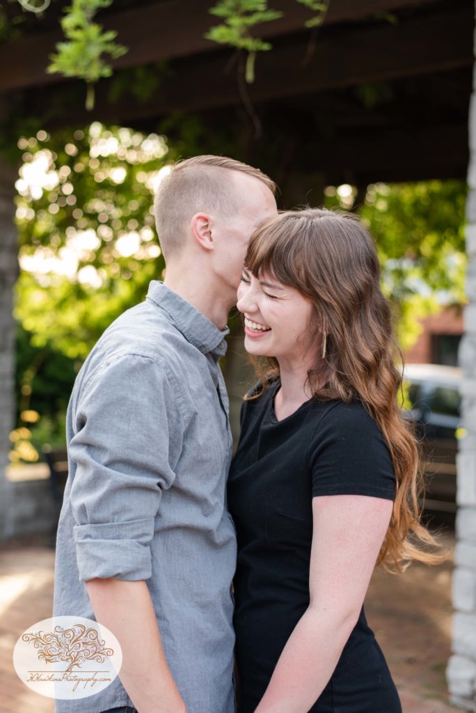 Groom to be whispers something in bride's ear during their engagement pictures at Franklin Square Park in Syracuse, NY