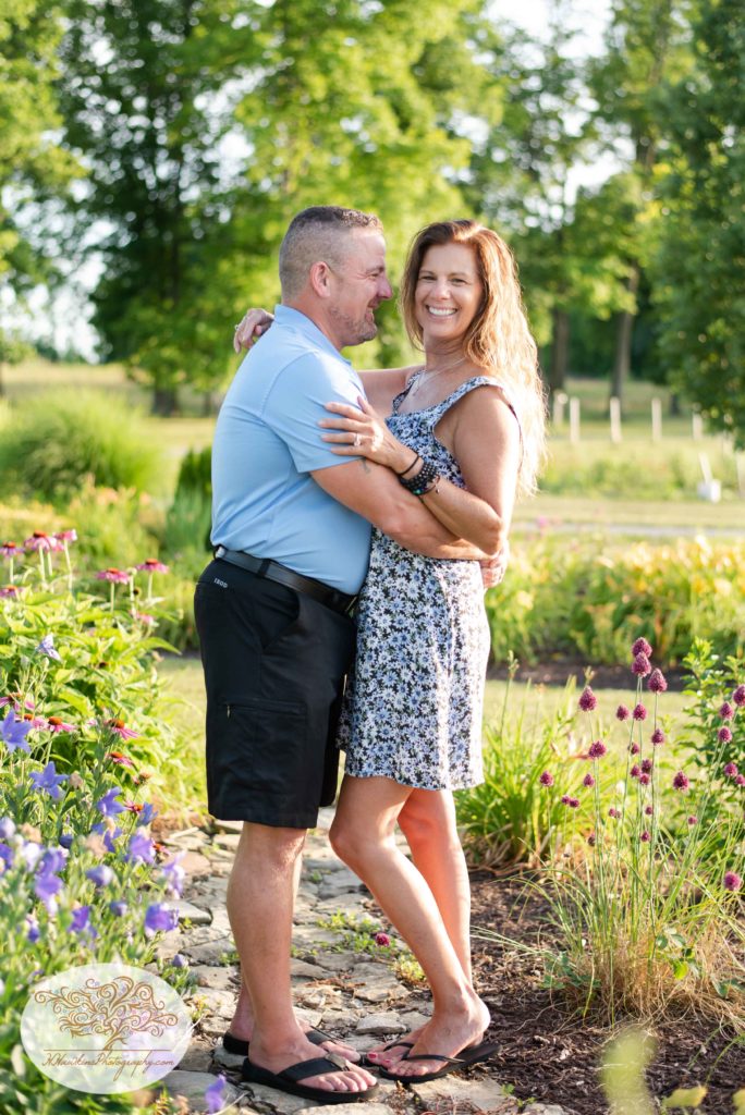 BRide to be looks at camera while groom nuzzles in standing in flower garden at Anyela's Vineyard in Skaneateles NY