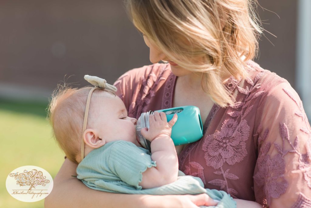 Baby drinks from bottle during summer family pictures while mom in a mauve dress holds her