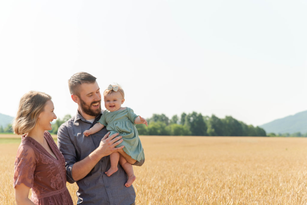 Mom, dad and baby laugh together while standing in a wheat field