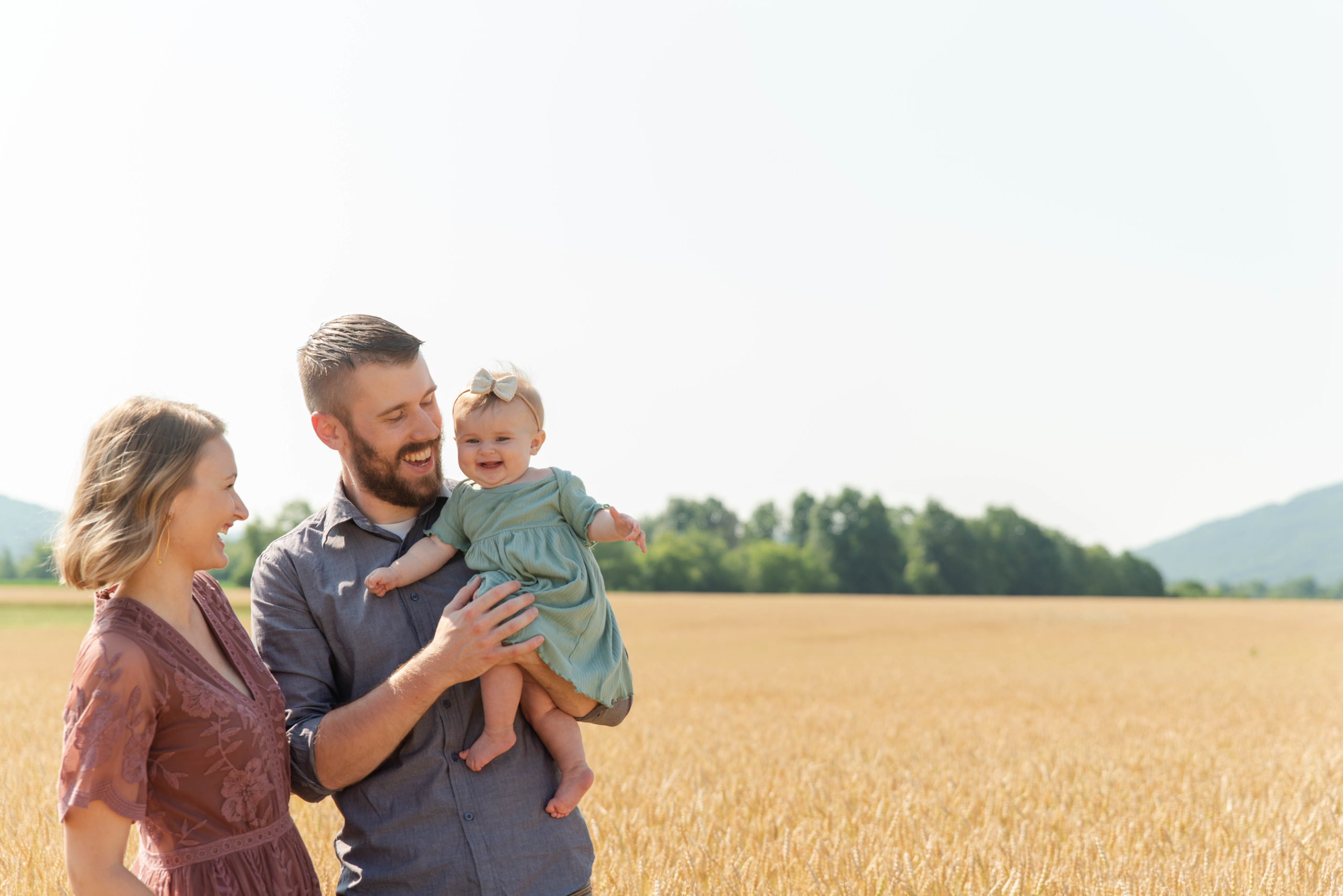 Mom, dad and baby laugh together while standing in a wheat field
