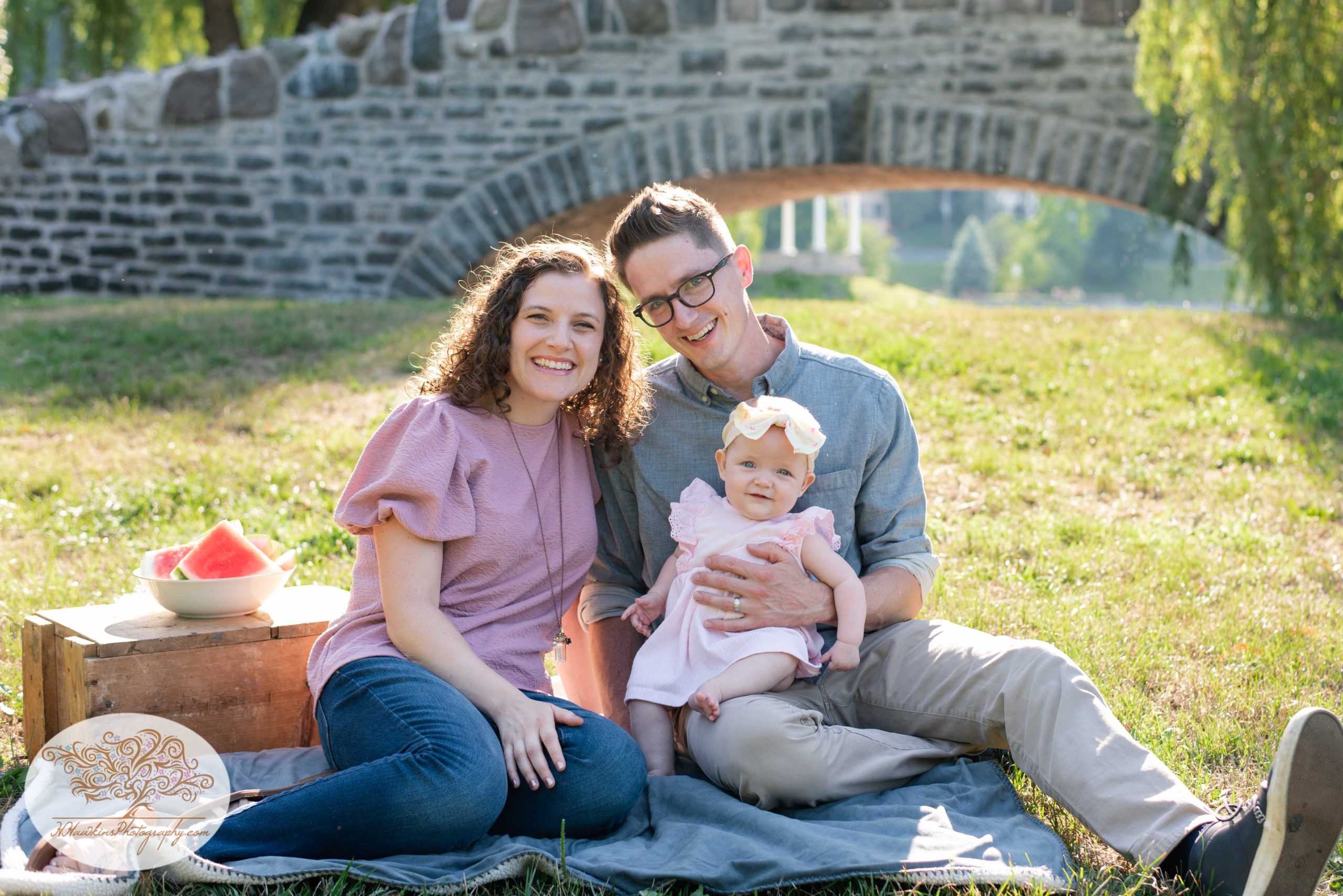 Mom, dad and baby girl in pink dress get their picture taken by Syracuse family photographer in front of the bridge at Upper Onondaga Park in Syracuse NY