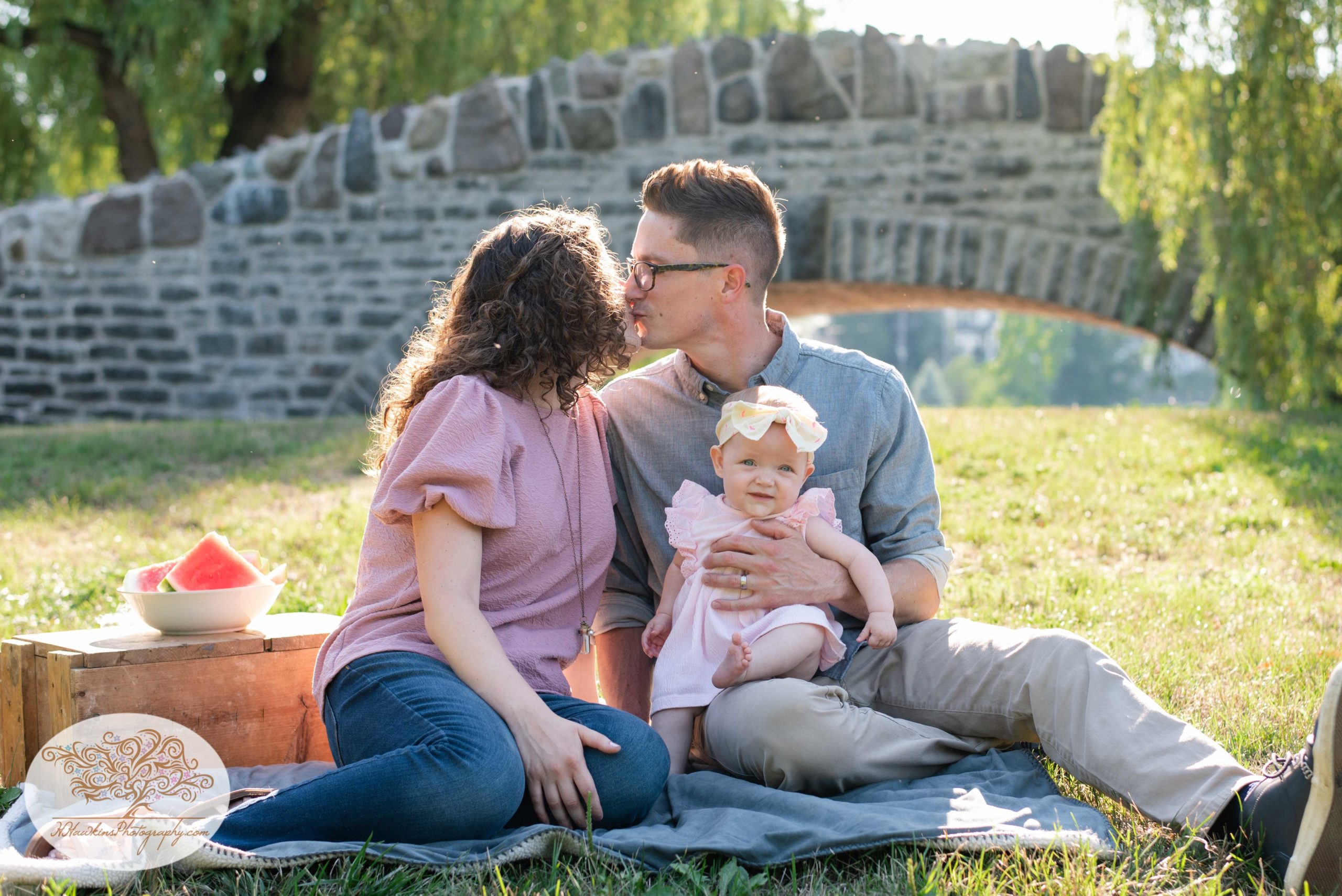 Mom, dad and baby girl in pink dress get their picture taken by Syracuse family photographer in front of the bridge at Upper Onondaga Park in Syracuse NY