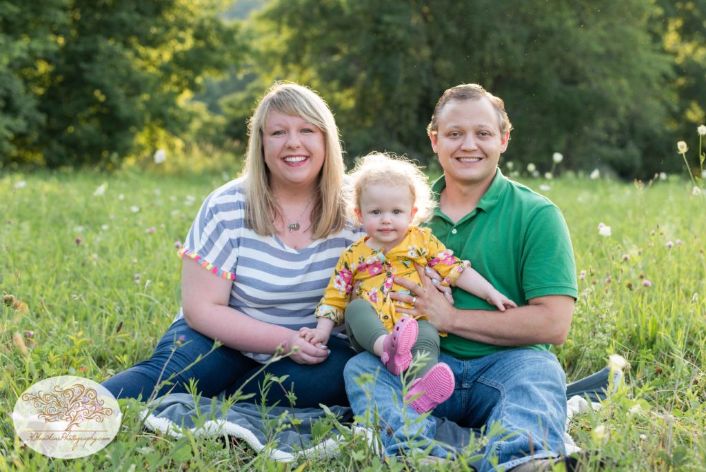 Mom dad and little girl smile for the camera sitting on the ground in a summer field of flowers for family pictures 