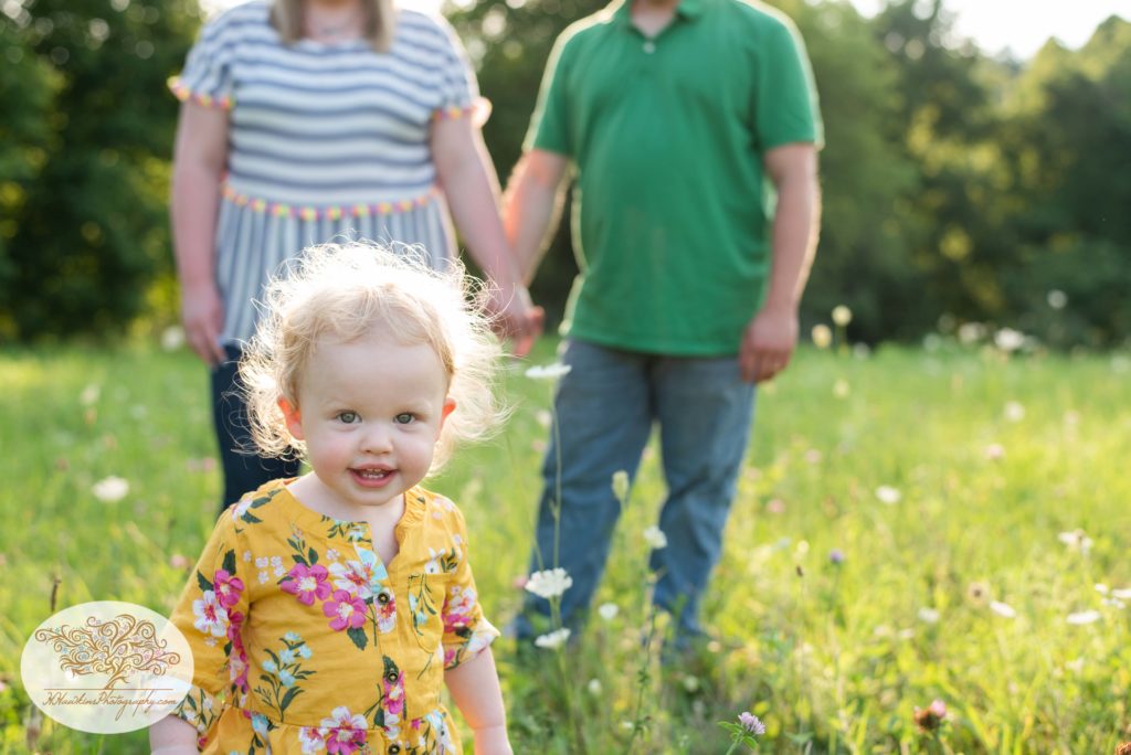 Toddler with curly hair smiles for camera while mom and dad stand in the background holding hands