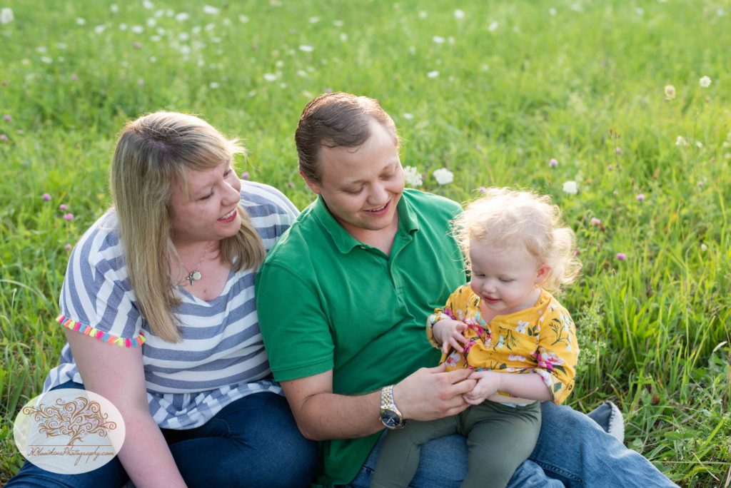 Mom, dad and little girl sit in a field of flowers for their summer family pictures