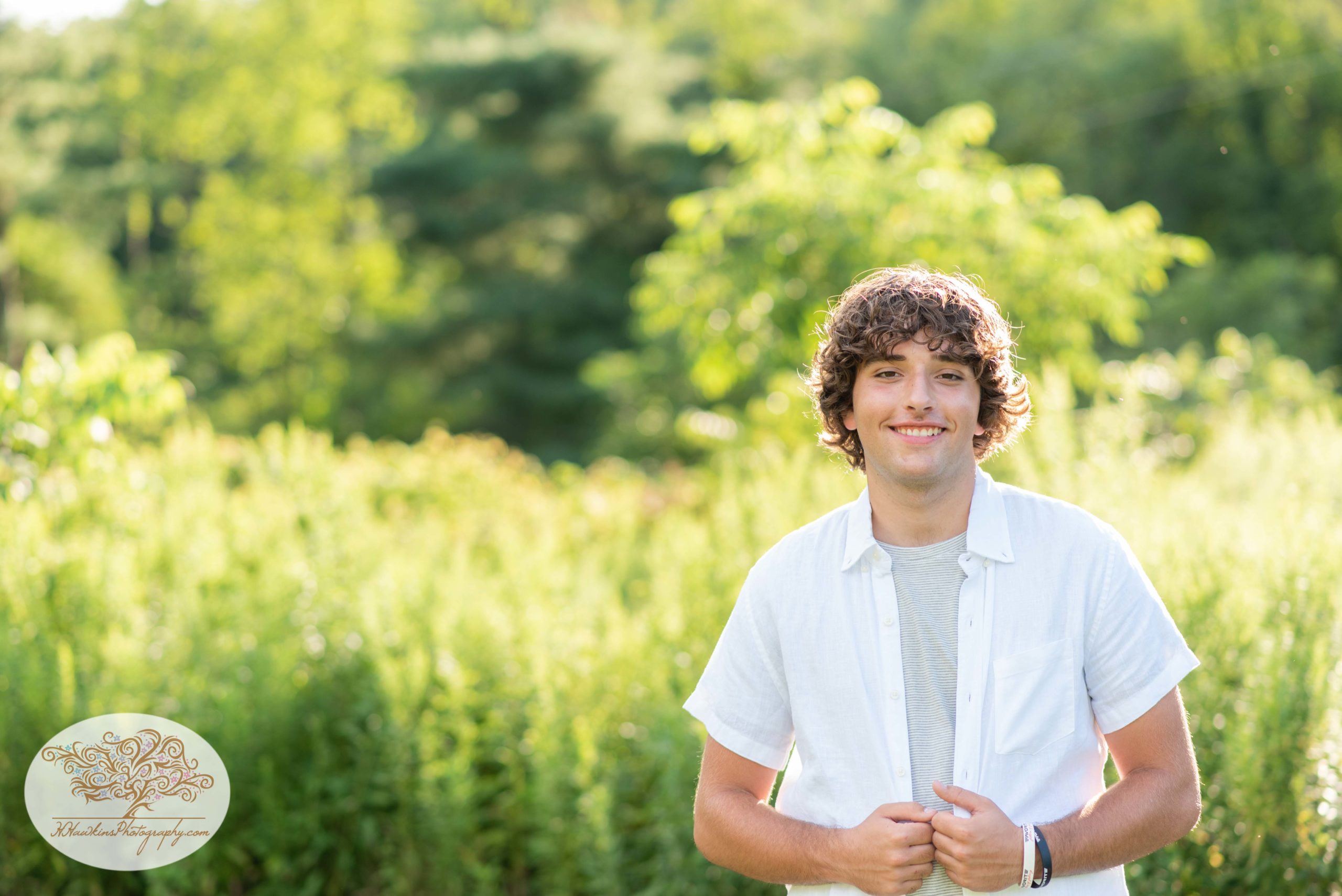High school senior guy holds his shirt in front as he stands in a sunlit field for his photo session