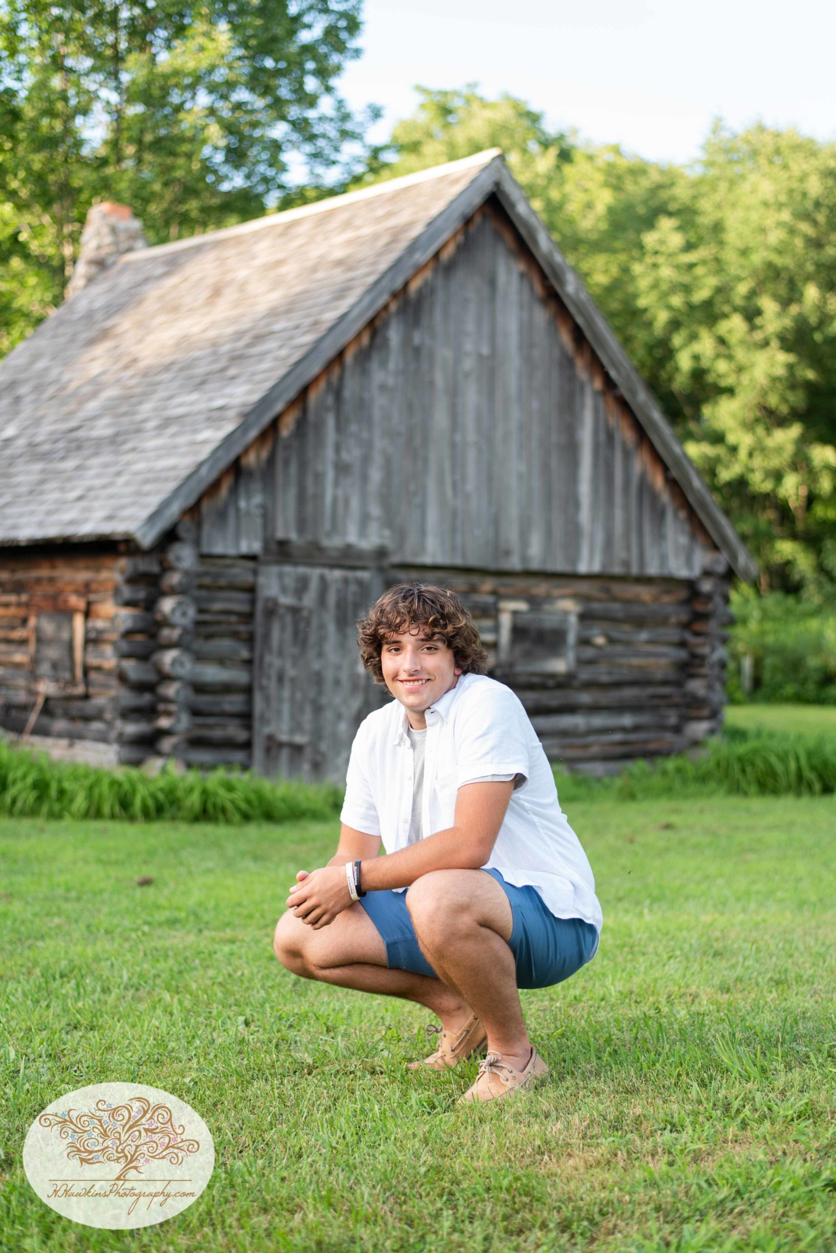 Syracuse high school senior smiles as he squats in front of log cabin at Baltimore Woods during his pictures