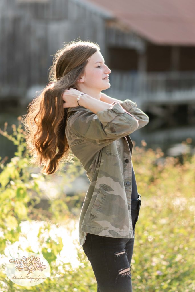 High school senior girl fixes her hair in ripped black jeans and a camo jacket with the sunshine behind her and a cabin in the distance at Camillus Erie Canal Park