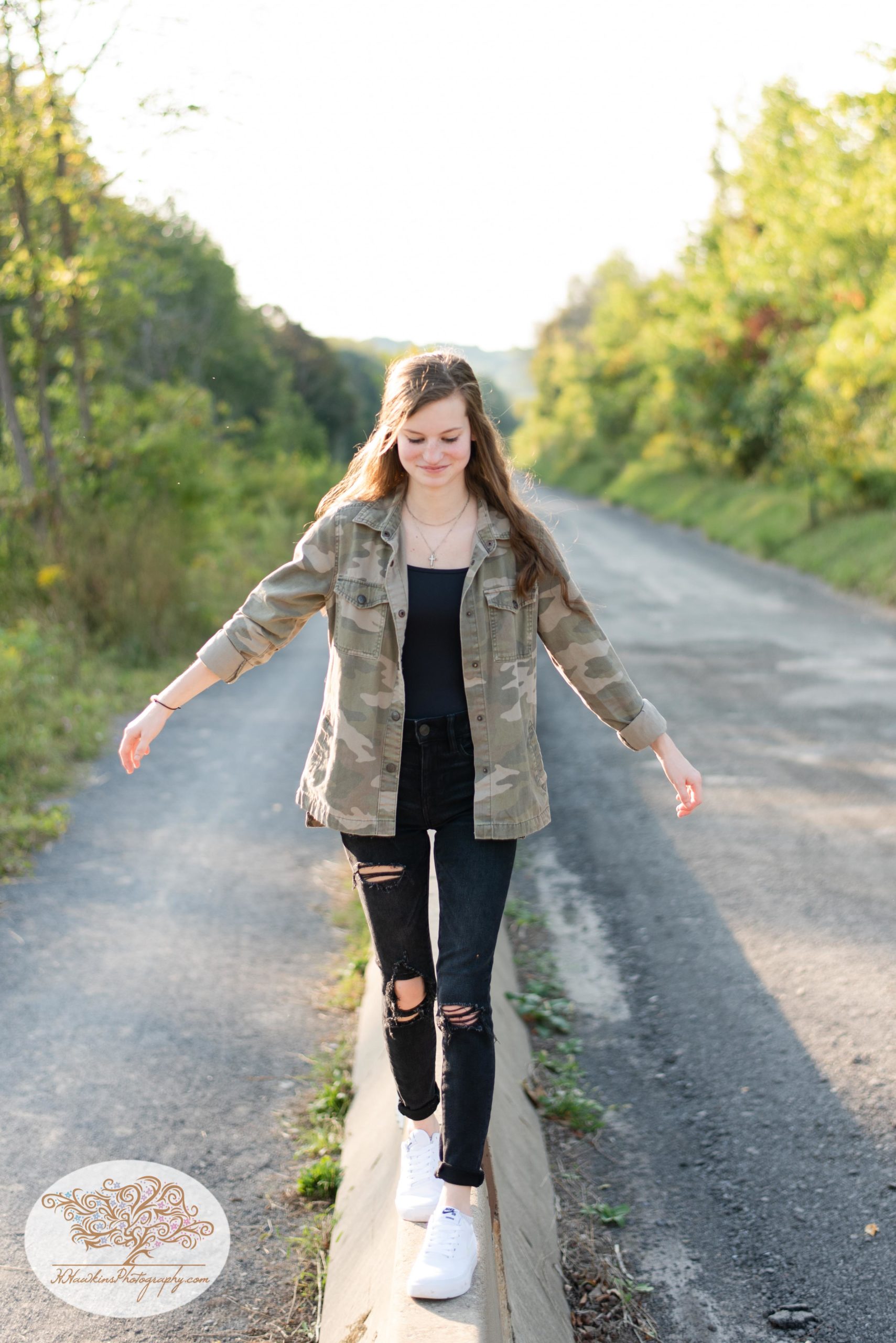 High school senior girl stands on concrete barrier near the Erie Canal in Camillus NY in black jeans and a camo jacket