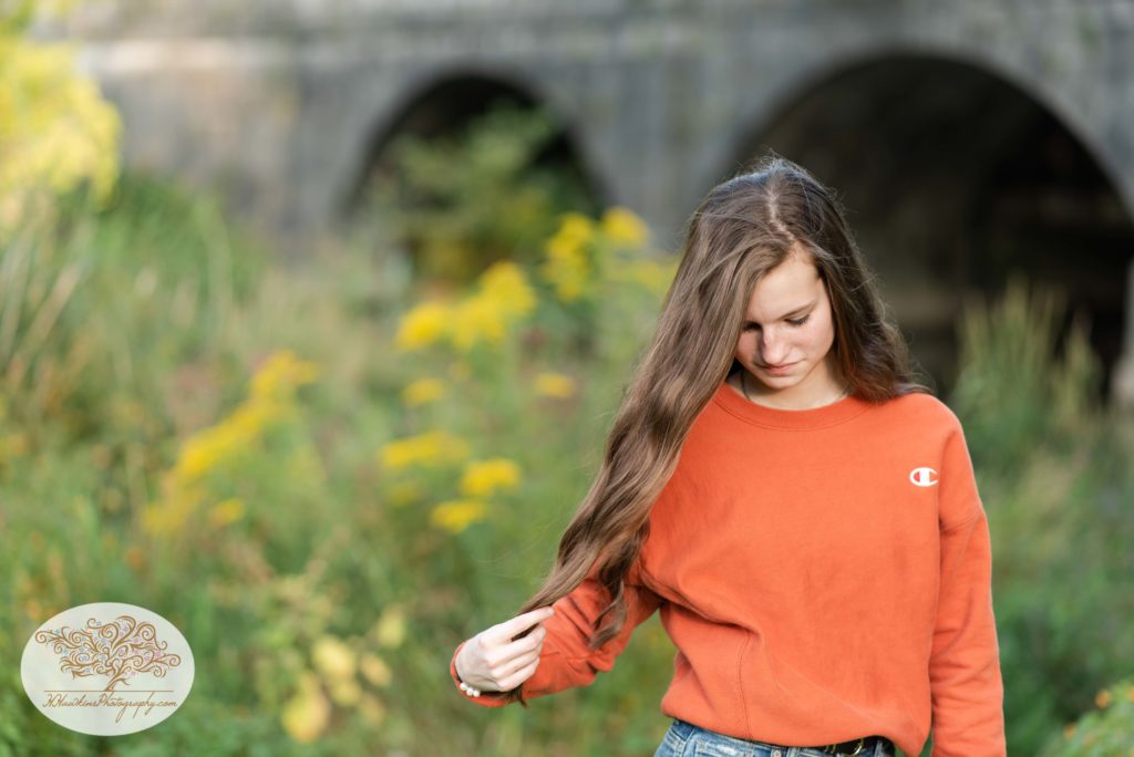 High school senior girl in blue jeans and an orange sweatshirt stands in front of Erie Canal aqueduct in Camillus NY 