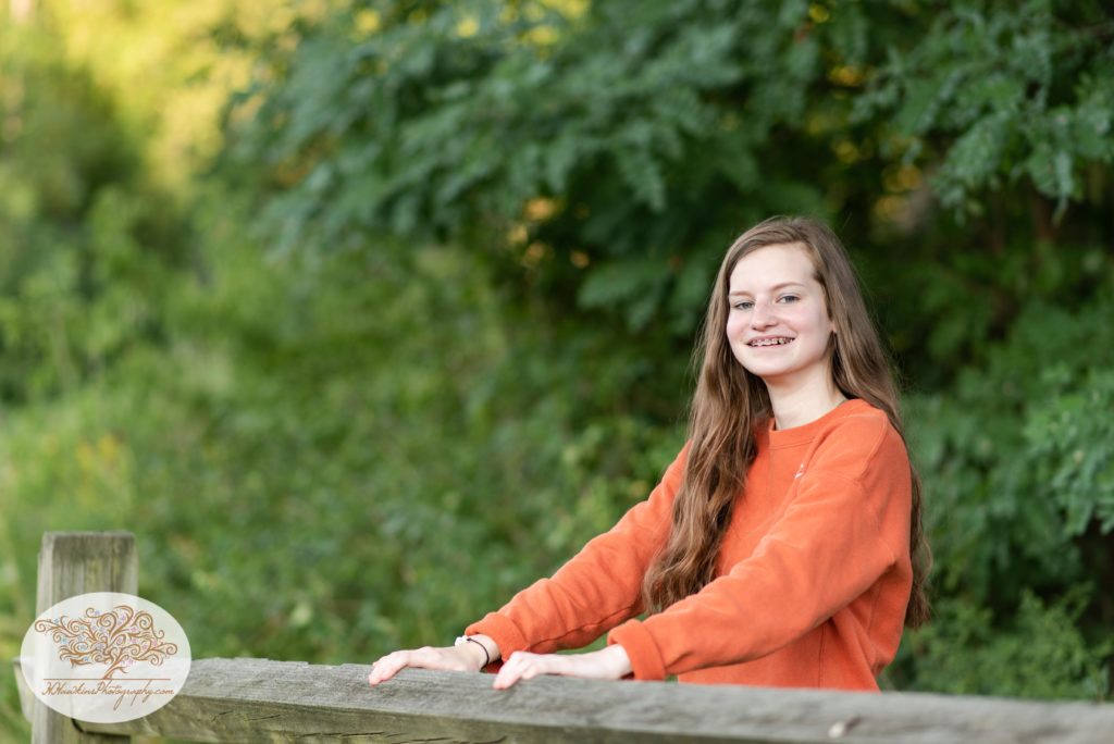 High school senior girl in blue jeans and an orange sweatshirt stands beside a fence in the field near the Erie Canal in Camillus NY