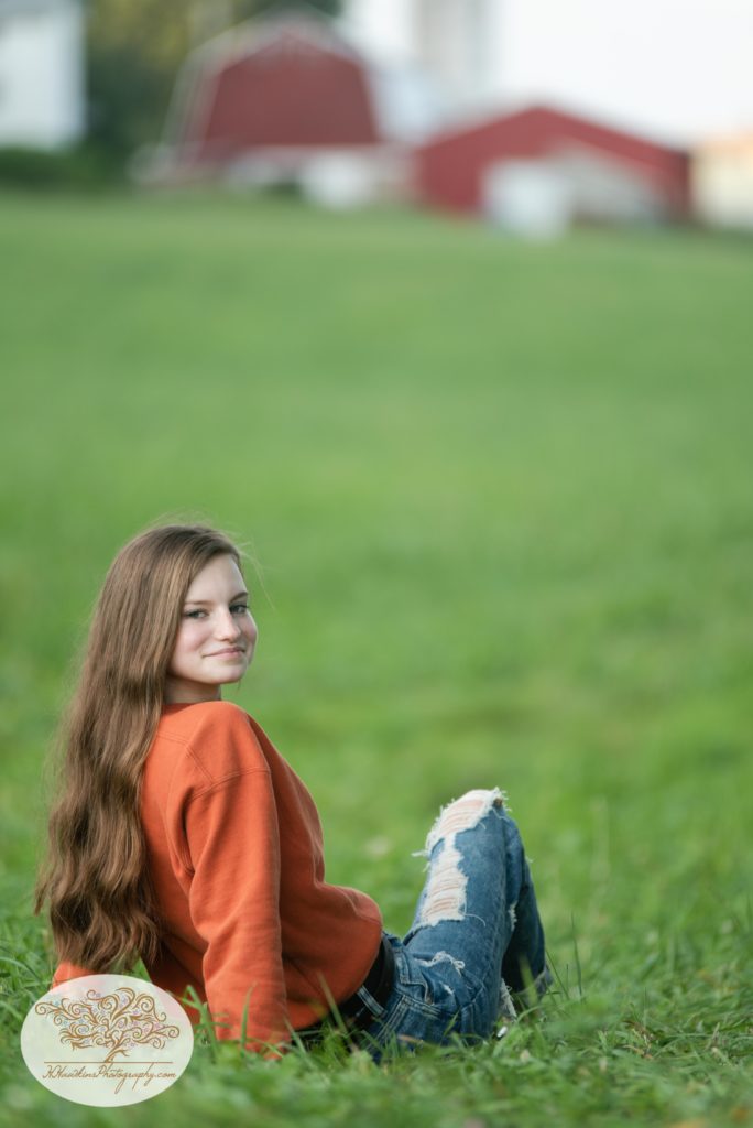 High school senior girl in blue jeans and an orange sweatshirt sits in a field front of a barn near the Erie Canal in Camillus NY