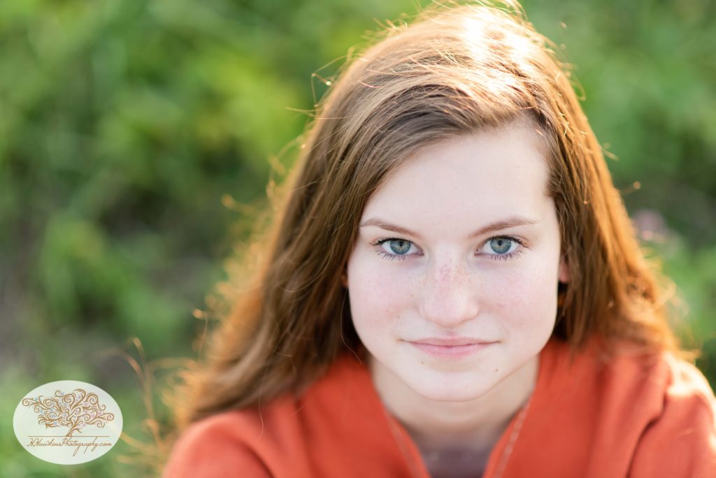 High school senior girl looks right into the camera lens with her green eyes and orange sweatshirt